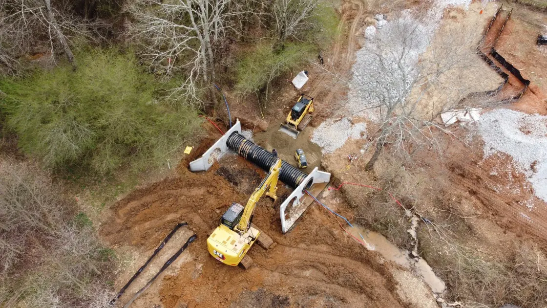 Aerial view of an excavator setting a large culvert pipe at a crossing