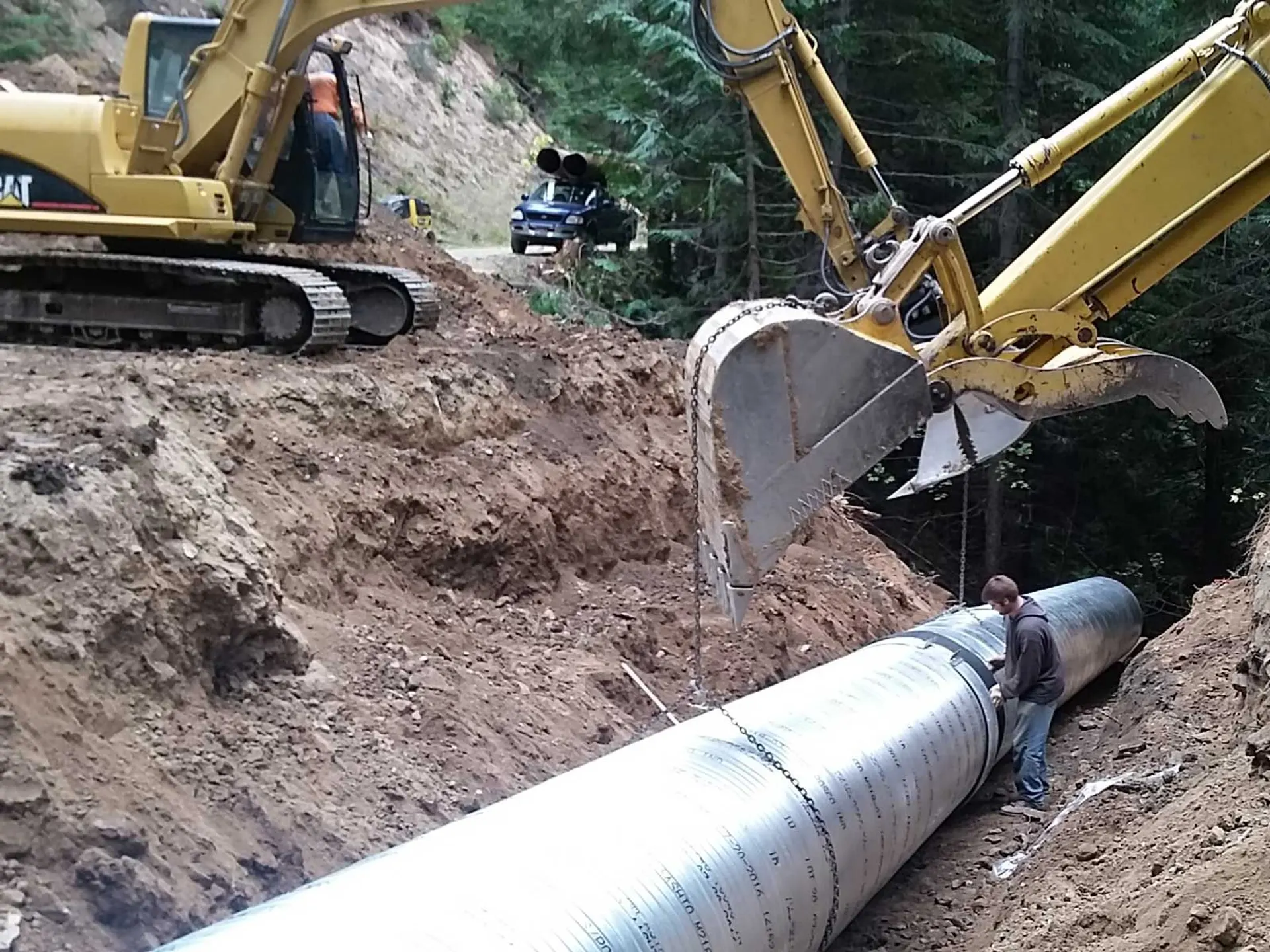 Excavator setting a large metal culvert pipe into a trench